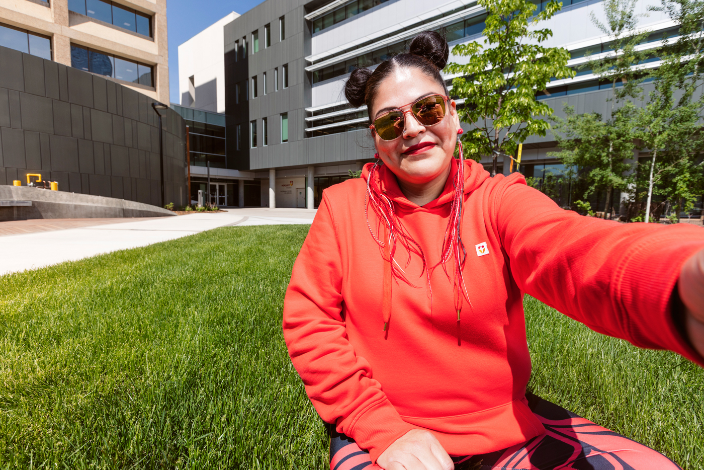 A female student sitting on the grass in the NorQuest greenspace taking a selfie