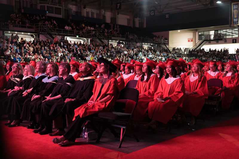 Group of diverse students in red robes sitting watching stage